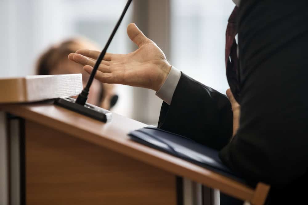 A person in a suit speaks at a podium with a microphone, gesturing with one hand. The background is out of focus, showing another person seated and slightly blurred. It appears to be an important event, perhaps featuring a renowned Criminal Defense Attorney from Cecil County.