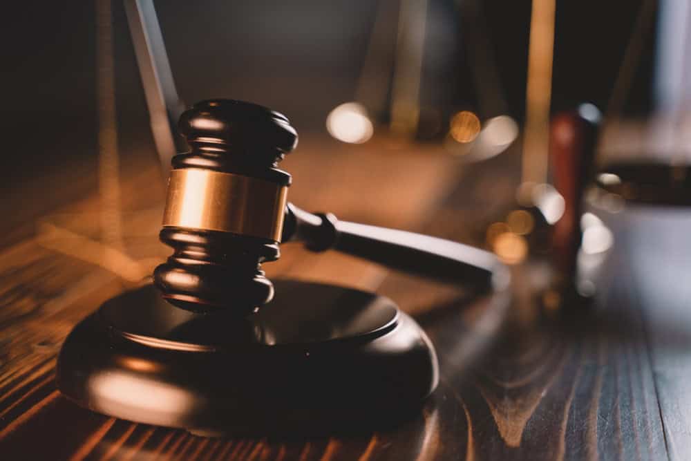 A close-up of a wooden gavel resting on its base, with blurred balance scales in the background on a wooden surface, symbolizing law and justice. The lighting is soft, casting reflections on the polished gavel and creating a warm ambiance.